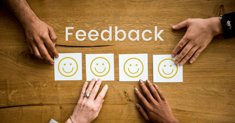 Ouça o feedback de clientes e colaboradores Mesa com mão de 4 pessoas diferentes, no meio 4 papéis com rostos felizes e a palavra Feedback escrita em cima
