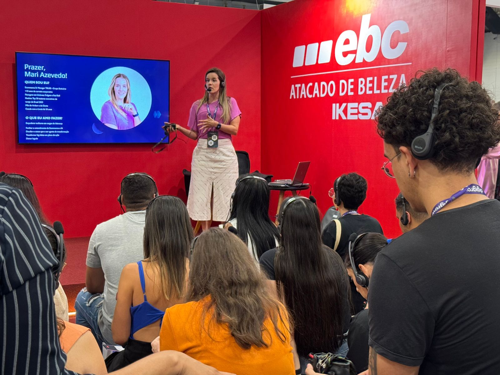 O que é o Mercado Livre Experience? Estande vermelho com uma palestrante loira de camiseta amarela e calça branca, uma televisão 80 polegadas com o fundo vermelho a foto e o texto da apresentação, pessoas com headset assistindo a palestra