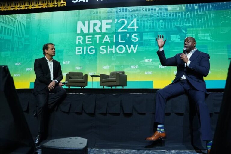 Futuro do Varejo de Beleza - Veja 7 insights da NRF. Ao lado esquerdo o apresentador, um homem branco de terno e gravata, ao lado esquerdo a lenda do basquete Magic Johnson também de terno e gravata, ao fundo um telão com fundo verde escrito NRF